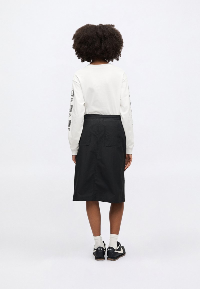 Black knee-length skirt with back pockets, worn over a white long-sleeve top featuring black graphic prints on sleeves.