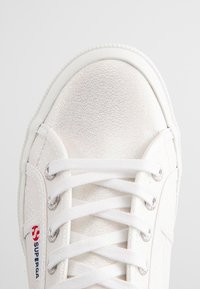 Sneaker bianca con una parte superiore testurizzata e iridescente. Presenta una suola in gomma bianca, occhielli e lacci argentati, con un accento del logo rosso di lato.