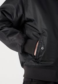 Main dans la poche d'une veste noire avec poignets et taille côtelés, montrant le détail du bouton-pression sur le rabat de poche.