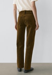 Person iført brune manchesterbukser med lige ben, hvid kortærmet top og sorte slip-on sko, stående mod en ensartet lys baggrund.