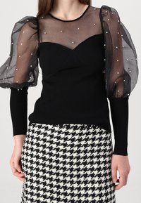 Svart topp med genomskinliga, puffärmar prydda med vita pärlor, i kombination med en kjol med svart och vit houndstooth-mönster.