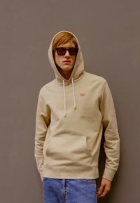 Beige hettegenser med snorer og frontlomme, med en liten rød logo. Kombinert med blå denimjeans og mørke solbriller.