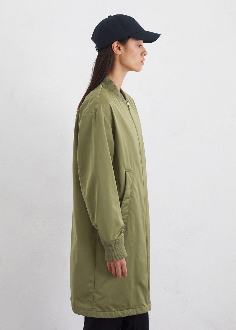 Olivgrüne Bomberjacke mit Reißverschluss, gerippten Bündchen und Seitentaschen, getragen mit einer schwarzen Kappe, die eine lässige Silhouette bietet.