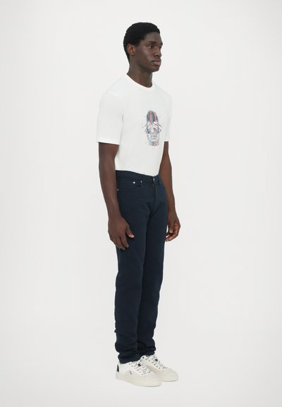 Camiseta blanca con un gráfico de calavera colorido, combinada con jeans azul oscuro y zapatillas blancas con detalles negros.