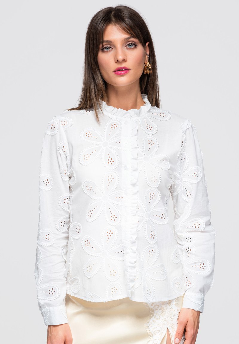 Femme portant un chemisier blanc à manches longues avec de grandes découpes en dentelle florale et un col à volants, associé à une jupe crème avec des détails en dentelle.