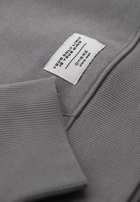 Tissu gris côtelé avec poignet et étiquette rectangulaire blanche portant l'inscription : « VOTRE SEULE LIMITE EST VOTRE ESPRIT », « OMBRE depuis 1995 ».