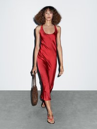 Robe maxi en satin rouge avec un décolleté arrondi, une silhouette ajustée et une longueur mi-mollet, associée à un sac tissé marron et des tongs roses.