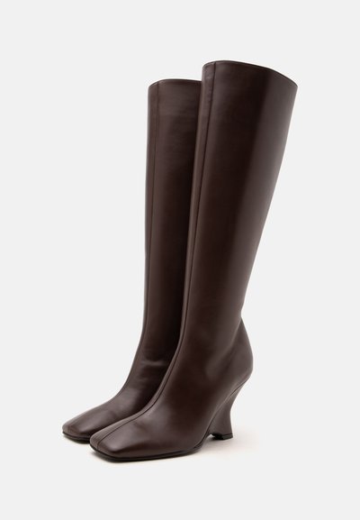 Paire de bottes hautes en cuir brun foncé avec talons bas inclinés et bouts carrés sur un fond clair uni.