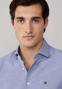 Giovane uomo con capelli corti e ricci di colore marrone, che indossa una camicia azzurra con bottoni e un piccolo logo ricamato sul petto.