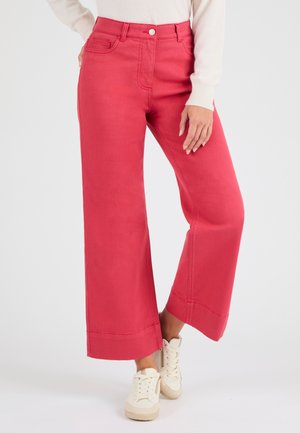 Vrouw in felroze, wijde, cropped broek en witte sneakers, staand tegen een effen witte achtergrond.
