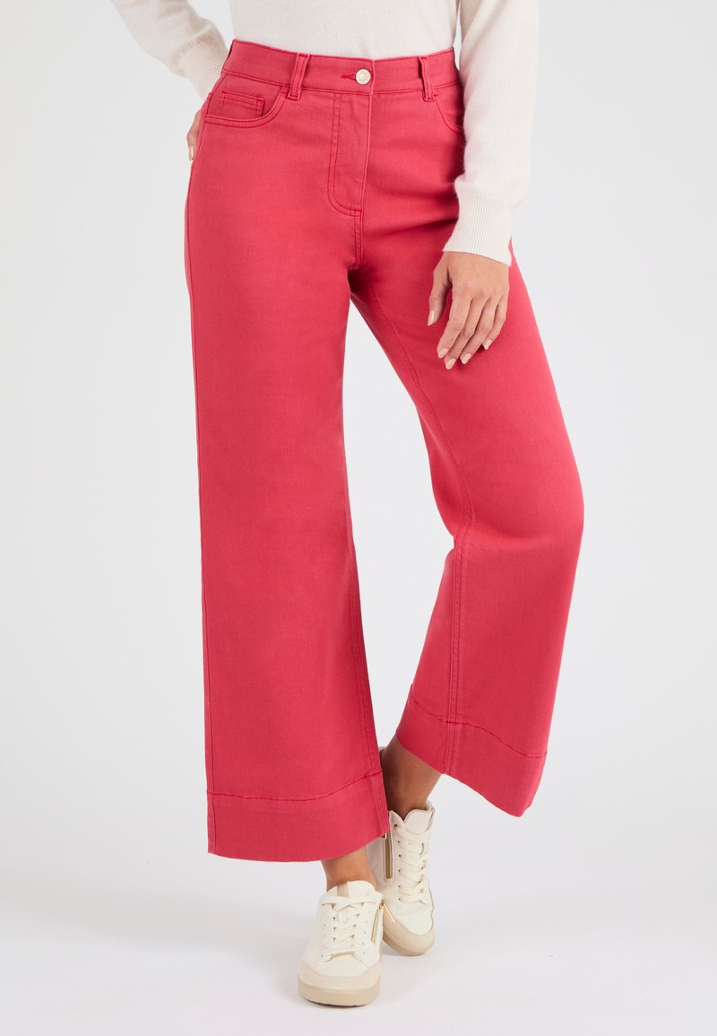 Mujer vestida con pantalones cropped de pierna ancha de color rosa brillante y zapatillas blancas, de pie frente a un fondo blanco liso.