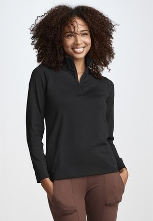 Lächelnde Frau mit lockigem Haar, die ein schwarzes, langärmliges Oberteil mit Reißverschluss und braune Hose trägt, Hände in den Taschen, vor einem weißen Hintergrund stehend.