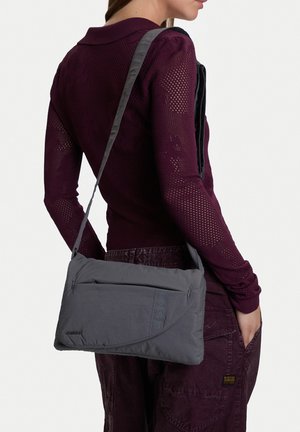 Kobieta ubrana w ciemnofioletowy strój niesie prostokątną, szarą torebkę na ramię z przednią kieszenią na zamek i długim paskiem przez ramię.