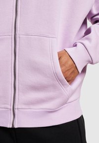Lys lilla zip-up hettegenser med frontlommer, ribbede mansjetter og nederkant, samt en glatt stoffoverflate. Glidelåsdetalj synlig.