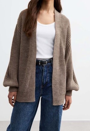 Vrouw draagt een losse beige gebreide cardigan over een wit hemdje en een hoge blauwe spijkerbroek met een zwarte riem, staand tegen een effen achtergrond.