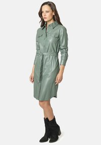 Vestido camisero de imitación de cuero verde con mangas largas, dos bolsillos en el pecho, un cinturón a la cintura y corte recto, combinado con botas negras hasta el tobillo.