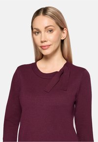 Bordowy sweter z dzianiny z okrągłym dekoltem i kokardą na lewym ramieniu. Gładka faktura z długimi rękawami.