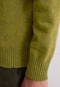 Grüner gestrickter Pullover mit einem strukturierten Rippenbund und -manschetten, verziert mit gelben und blauen Sprenkeln. Prägiertes Logo am unteren Rand.