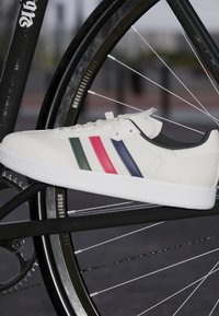 Białe sneakersy z teksturowaną cholewką, zdobione zielonymi, różowymi i granatowymi paskami. Biała gumowa podeszwa. Umieszczone na czarnej ramie roweru.