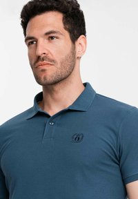 Polo tričko v tmavě modré látce s krátkými rukávy, límcovým výstřihem a dvěma knoflíky. Obsahuje vyšívané logo v oblasti hrudi.