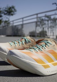 Lichtbeige hardloopschoenen met oranje strepen en blauwe veters, rustend op een geplaveide ondergrond nabij een metalen reling buiten.
