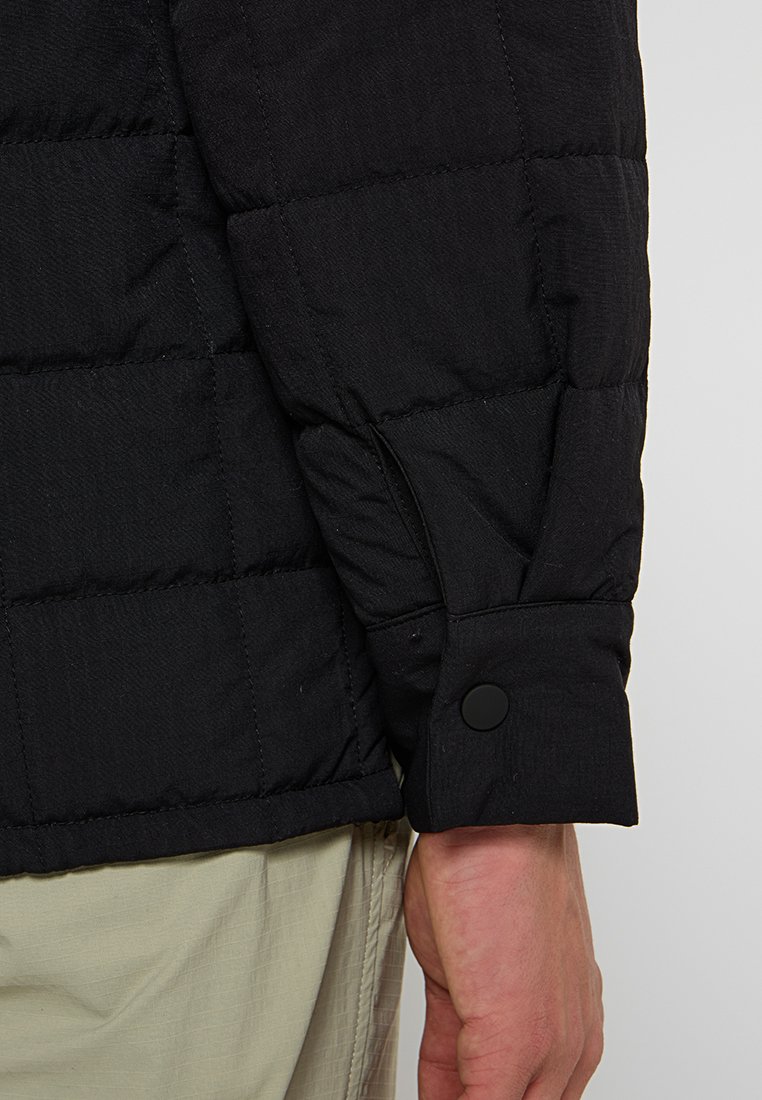 Svart vadderad jackärm med kviltad struktur, med en rund svart knappstängning och en något avsmalnande manschettdesign.