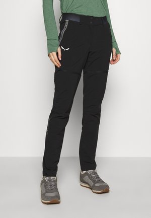 Person iført sorte udendørsbukser med lynlåslommer, grå sneakers og en grøn langærmet top, stående på en ensartet lys baggrund.