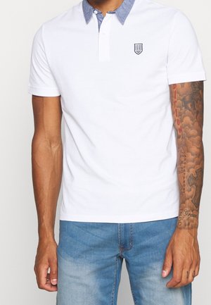 Polo blanc avec un col en chambray, manches courtes, et un petit logo brodé sur la poitrine. Porté avec un jean denim bleu clair.