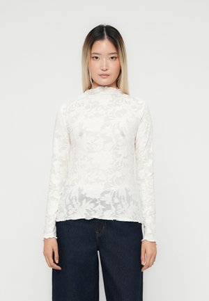 Femme aux cheveux raides du noir au blond portant un haut en dentelle florale blanche à manches longues et un pantalon large taille haute foncé, debout devant un fond uni.