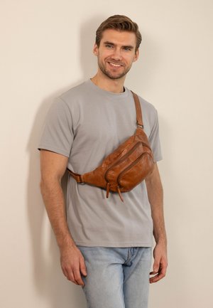 Uomo sorridente che indossa una t-shirt grigia e jeans azzurri chiari con una borsa a tracolla in pelle marrone davanti a una parete beige.