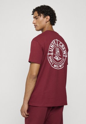 Homme aux cheveux bouclés portant un t-shirt bordeaux et un pantalon assorti, montrant un graphique circulaire blanc et un texte au dos du t-shirt.