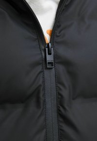 Veste rembourrée noire avec une fermeture éclair centrale, texture lisse et une accent orange bien visible. La tirette de la fermeture éclair affiche un logo de la marque.
