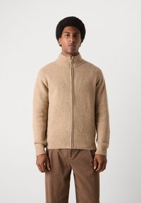 Beige zip-up genser med høy krage, ribbet tekstur og lange ermer. Bært med brune bukser, presentert mot en ensfarget bakgrunn.