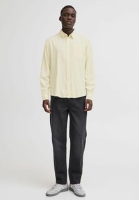 Lys gul skjorte med knapp foran og brystlomme, kombinert med mørke rette jeans og grå sneakers. Jevn tekstur og avslappet passform.