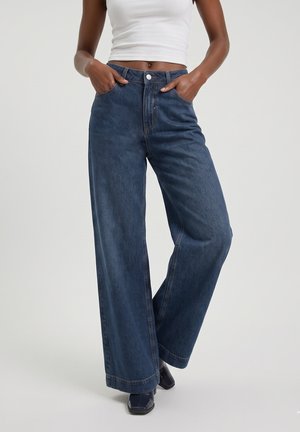 Femme portant un jean bleu foncé taille haute à jambes larges, un débardeur court blanc et des chaussures noires à bout carré, mains dans les poches.