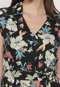 Mujer vestida con un vestido negro de flores grandes en blanco, rojo y azul, con botones en el frente, cuello y un lazo en la cintura.
