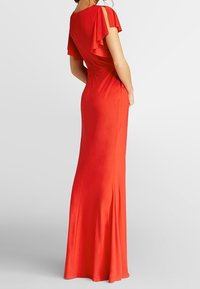 Femme portant une robe rouge longue avec des manches volantées et des fentes, se tenant avec le dos partiellement tourné sur un fond blanc.