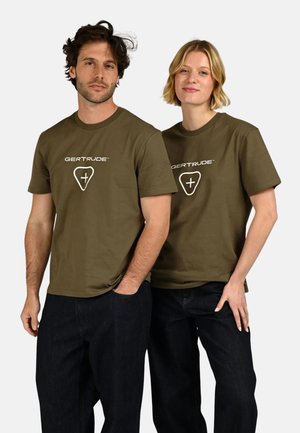 Homme et femme portant des t-shirts assortis « GERTRUDE » vert olive et des pantalons foncés, debout de manière décontractée devant un fond blanc uni.
