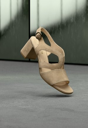 Beige suède sandaal met hoge hak, open teen, uitgesneden zijkanten en een ronde gesp op de enkelband.