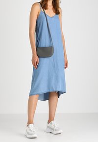 Abito in denim azzurro chiaro con scollatura a V, spalline sottili e un leggero orlo asimmetrico. Abbinato a sneakers bianche e a una piccola borsa a tracolla grigia.