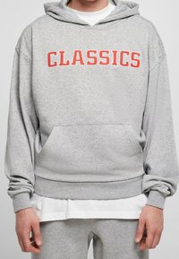 Grijze hoodie met rode tekst "CLASSICS" op de borst. Voorzien van een kangoeroezak en geribbelde manchetten aan mouwen en zoom. Zachte stof.