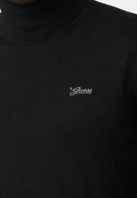 Gros plan d'un pull col roulé noir avec le logo "Guess" blanc brodé sur le côté gauche de la poitrine.