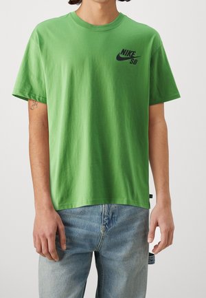 Osoba ubrana w zielony t-shirt Nike SB i niebieskie dżinsy, stoi z rozluźnionymi ramionami wzdłuż ciała.