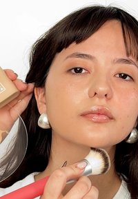 Un lápiz de maquillaje beige en una mano, un pendiente de aro plateado y un pincel de maquillaje con un mango rojo en la otra mano, contra un fondo blanco.