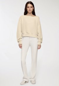 Felpa beige cropped con ampio scollo a barca, polsini e orlo a coste; abbinata a jeans bianchi a vita alta e sneakers in tonalità neutre.