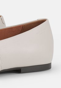 Scarpa in pelle grigia chiara con tacco basso e curvo e fodera interna marrone. Presenta un design minimalista e una piccola suola nera.