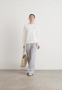 Felpa bianca, pantaloni larghi grigi, sneakers argentate, tiene una borsa a tracolla beige. Tessuto morbido, vestibilità rilassata, design minimalista.