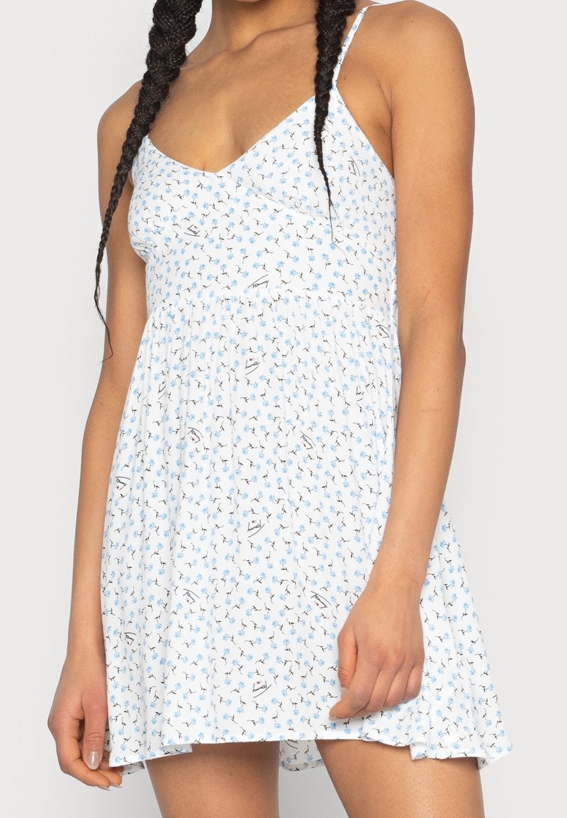 Vit klänning med blå blommönster, V-ringning, tunna justerbara axelband, flödande A-linjeform och lättviktsmaterial.