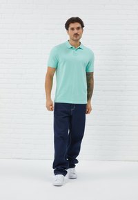Polo shirt bleu-vert clair avec col et manches courtes, associé à un jean en denim bleu foncé et des baskets blanches, devant un mur de briques blanches.