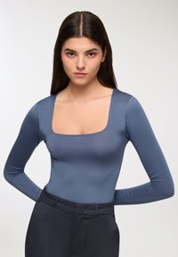 Bodysuit z długim rękawem w odcieniu niebiesko-szarym, z kwadratowym dekoltem i dopasowanym krojem. Gładka tkanina o lekko błyszczącej powierzchni. Połączony z ciemnymi spodniami.